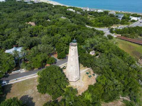 Bald Head Island, United States - 17 June 2023: Aerial view of Old Baldy Lighthouse and Smith Island Museum surrounded by dense maritime forest and coastal scenery.