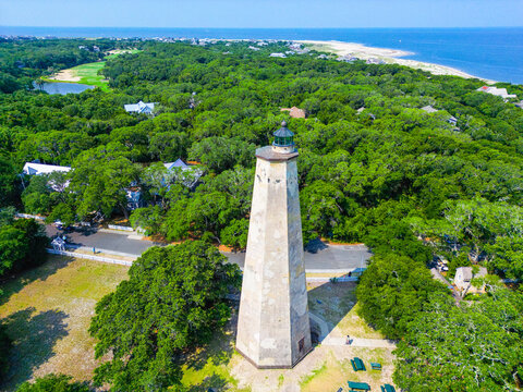 Bald Head Island, North Carolina, United States - 17 June 2023: Aerial view of Old Baldy Lighthouse and Smith Island Museum surrounded by lush maritime forest and coastal vegetation.