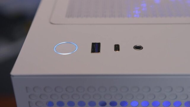 Low-angle close-up of desktop computer case top panel with glowing blue power button and USB ports showing illuminated power detail, brushed metal surface, and audio jack