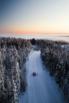 Aerial view of a Range Rover driving on a snow-covered road through a dense pine forest during a colorful winter sunset in Sirkka, Lapland, Finland.