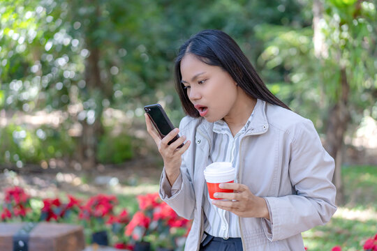 A surprised young Asian woman looking at her mobile phone with a shocked expression while holding a red takeaway coffee cup outdoors in a scenic park surrounded by lush green foliage and red flowers