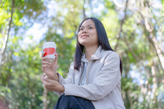 A smiling young Asian woman looking up thoughtfully with a gentle expression while holding a red takeaway coffee cup outdoors in a scenic green forest park surrounded by lush trees and natural light.