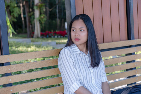 A thoughtful young Asian woman sitting on a wooden park bench outdoors, wearing a stylish striped shirt and looking away pensively with a calm expression against a beautiful background of green trees.