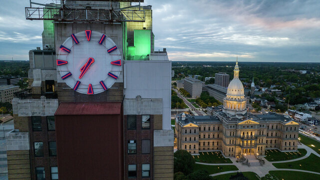 Lansing, United States - 23 September 2022: Aerial view of the Michigan State Capitol illuminated at dusk, next to a historic clock tower with red and blue neon lighting under a cloudy sky.