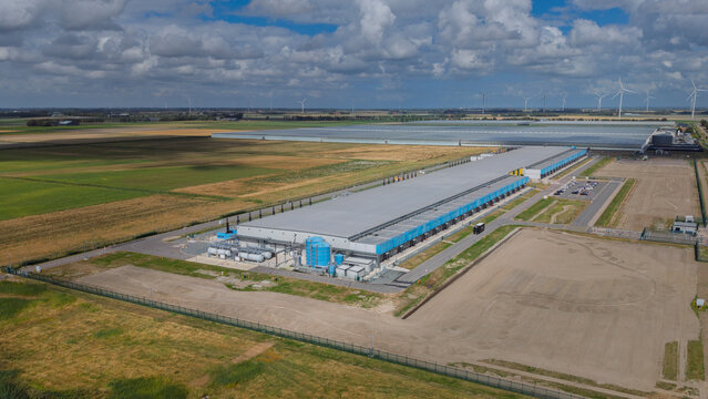 Aerial view of the Google data center Agriport A7 featuring large industrial buildings, blue cooling tanks, and surrounding agricultural landscape in Middenmeer, North Holland, Netherlands.
