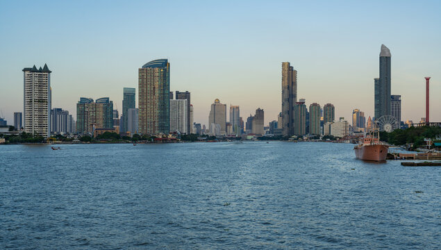 View of the Bangkok skyline along the Chao Phraya River featuring modern skyscrapers, a cargo ship moored near the bank, and a clear sky at dusk in Bangkok, Thailand.