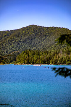 View of Eibsee lake with people paddleboarding on the turquoise water surrounded by dense pine forests and mountains in Grainau, Bayern, Germany.