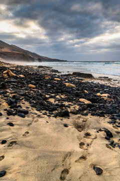 View of Playa de Cofete with dark volcanic rocks scattered across the sandy beach, ocean waves crashing on the shore, and mountains under a cloudy sky in Canary Islands, Spain.