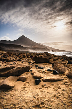 View of Playa de Cofete with rugged volcanic rocks scattered across the sandy beach, dramatic mountains rising in the background under a cloudy sky in Canary Islands, Spain.
