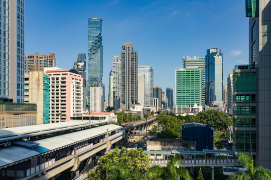 View of the Bangkok skyline featuring the iconic MahaNakhon skyscraper, modern office buildings, and the elevated BTS Skytrain tracks under a clear blue sky in Bangkok, Thailand.