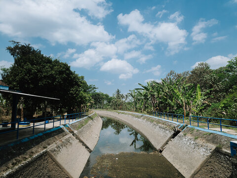 View of the concrete-lined irrigation canals of the Talangagung Metro Siphon structure, surrounded by lush vegetation and trees under a bright blue sky in Malang, East Java, Indonesia.