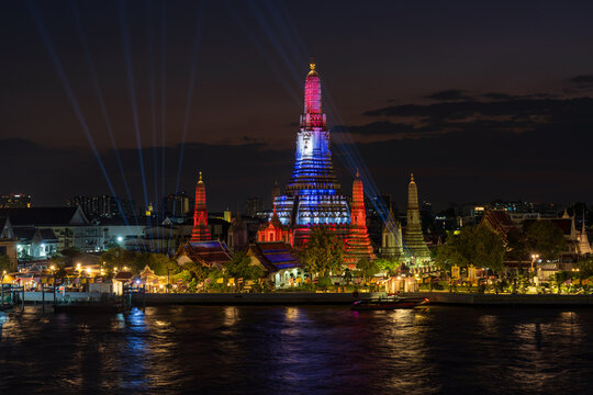 View of the illuminated Wat Arun temple at night with its central prang glowing against the dark sky and reflecting on the Chao Phraya River in Bangkok, Thailand.