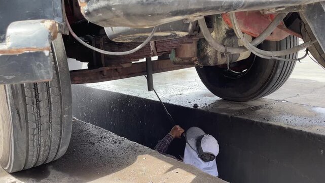 A worker in protective clothing pressure washes the underside of a heavy truck, directing a powerful water spray stream toward the chassis, suspension, pipes, and mechanical components below.