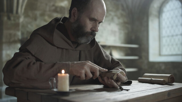 Bearded medieval scribe sharpening a penknife blade on a whetstone at a wooden table near a burning candle and scrolls inside a scriptorium