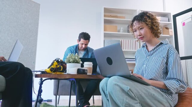 Side view of focused Asian female programmer sitting on floor cushion and typing software code on laptop.