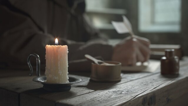 Lit candle on a metal holder on a rustic wooden table with a blurred monk writing with a quill pen in background inside a scriptorium