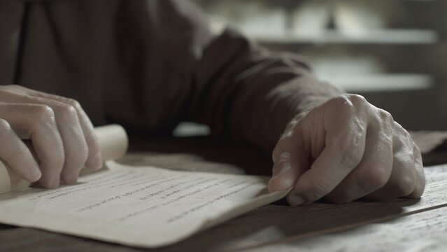 Hands of a medieval monk unrolling a parchment paper scroll with text on a wooden table inside a scriptorium