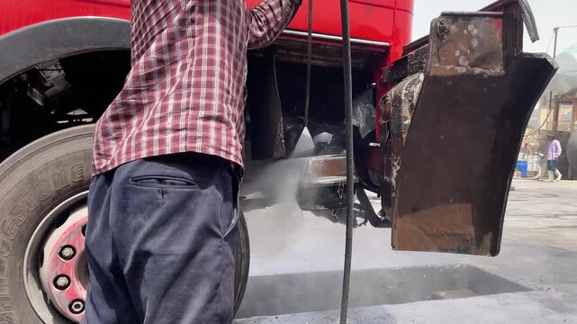 Close view of a worker pressure washing the underside of a red heavy truck, directing water toward the chassis, pipes, suspension, and lower mechanical parts covered with grime. onsite today