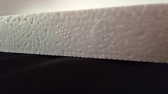 A horizontal macro view highlighting the edge and porous pattern of a rigid white expanded polystyrene board resting on black cloth under clear studio illumination. Great for technical visuals.