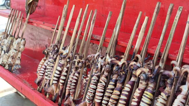 Camera pans up showing smoked squid skewered on bamboo sticks arranged neatly on a vibrant red wooden stall at a traditional coastal market.
