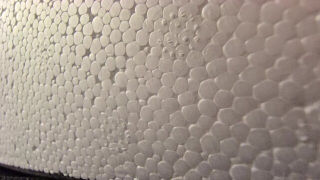 A close-up macro tracking shot over the bumpy, porous surface of white expanded polystyrene foam, highlighting its intricate cellular bead texture under warm studio side lighting.