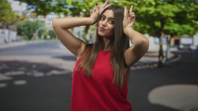 Young hispanic woman making bunny ears with hands and puckered lips on a tree lined urban street; playful.