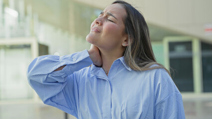 Obraz premium Young hispanic woman clutching neck and wincing while wearing a blue striped shirt outside a modern building; discomfort recovery.