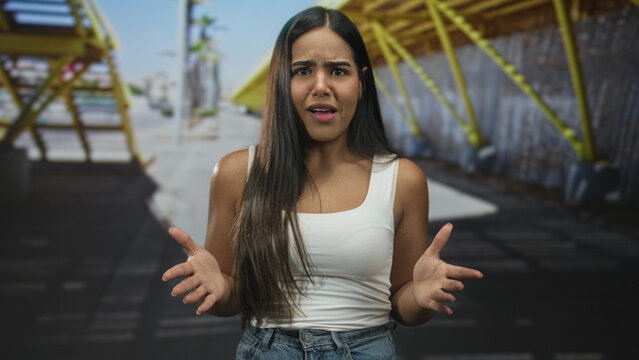 Hispanic woman with open palms and pleading expression gesturing toward camera on a street under yellow structure; frustration plea.
