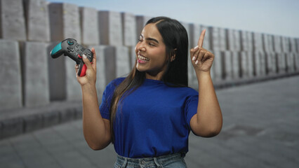Young woman holds a gamepad with one hand and points finger upward with the other in a concrete street setting lined by pillars while smiling  playful confidence. © Krakenimages.com