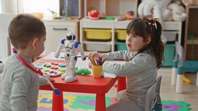 Little girl and boy playing with a toy robot and stacking rings at a red table in a building playroom; sibling bonding curiosity.