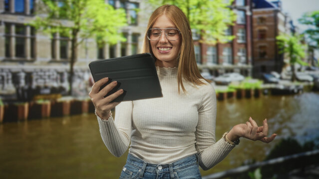 Woman redhead with glasses holding tablet and gesturing with hand on a canal street by water and buildings, wearing white top and jeans; curiosity.