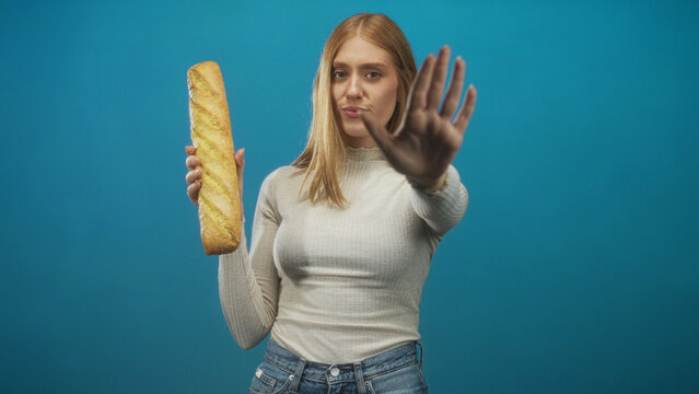 Young redhead woman holds baguette in left hand and shows palm out in stop gesture to camera in studio; rejection food refusal.