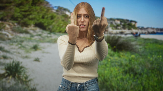Redhead woman shows both middle fingers to camera with tense hands at a beach shoreline, wearing cream sweater and jeans, stern expression; defiance.