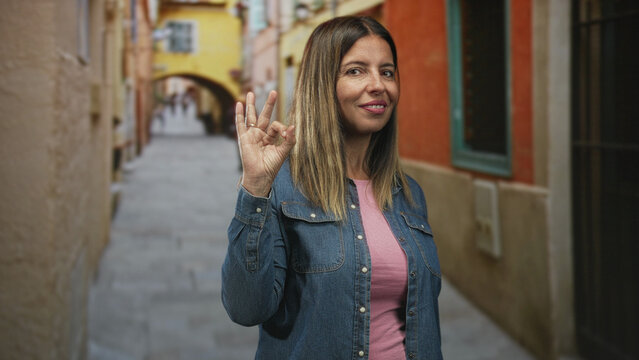 Woman shows ok sign with raised hand in a narrow street, wearing denim jacket and pink shirt, smiling at camera; approval contentment.