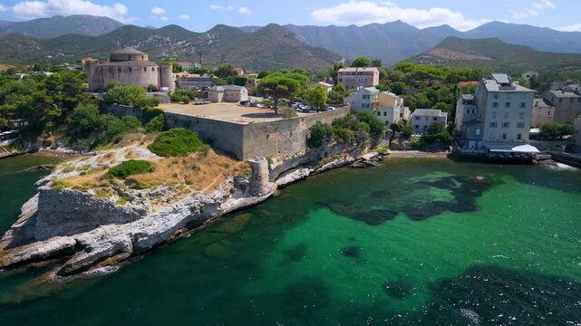 Saint Florent Citadel on rocky coast aerial drone rising view in Corsica France. Sunny blue sky turquoise sea and historic town turning to marina cinematic footage 4K