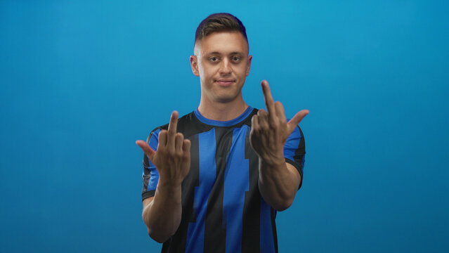 Young blond man in blue and black soccer jersey shows both middle fingers in a studio setting; defiance attitude.