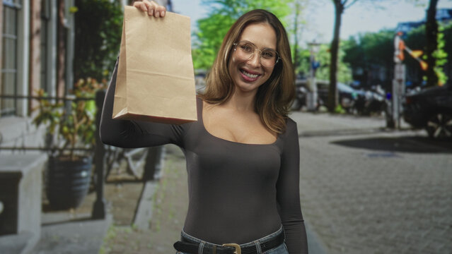 Woman holding paper bag up on street, smiling while wearing glasses and casual jeans with belt visible; friendly delivery service.