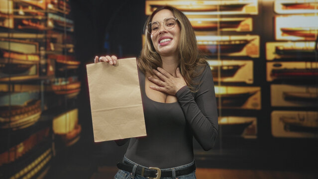 Woman holding paper bag, hand on chest in building while smiling and presenting a parcel; gratitude gift surprise.