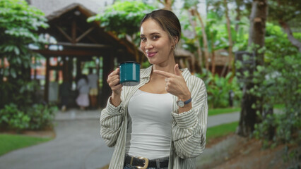 Fototapeta premium Woman holding teal mug and pointing at camera at building entrance, wearing white tank and striped shirt; confidence.