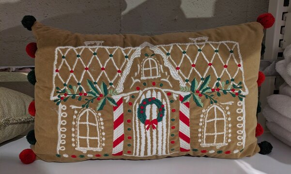 Tiempo de Navidad. Coj&iacute;n con dibujo de una casita de jengibre decorada con una guirnalda y hojas y bayas de acebo, en estante de tienda de decoraci&oacute;n