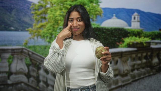 Woman holding custard apple and pointing to cheek on a stone terrace balustrade in building; thoughtful curiosity.