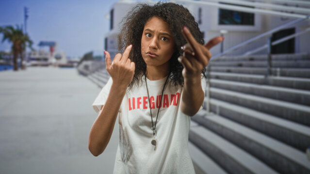 Woman lifeguard with curly hair flips both middle fingers with raised hands on seaside boardwalk steps; defiance.
