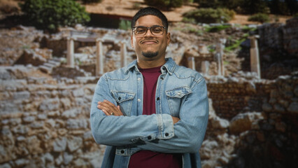 Fototapeta premium Hispanic man with beard and glasses adjusts frames and crosses arms in a studio in front of ancient stone ruins wearing denim jacket; confidence poise.