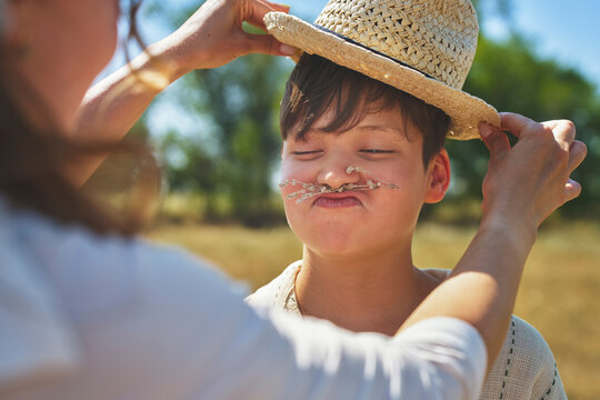 Mother putting straw hat on playful son making a funny face outdoors