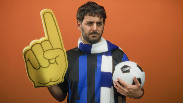 Man holds soccer ball and foam finger pointing up while wearing team scarf and jersey in studio; team pride.