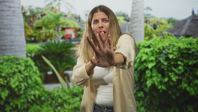 Woman covers face with hands and pushes palms forward in a tropical garden, making a stop gesture and hiding her expression; fear privacy.