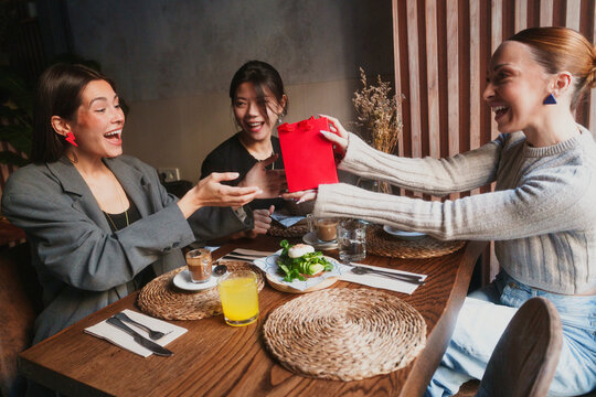 A woman gives a red gift bag to her surprised friend at a restaurant