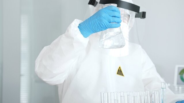 Scientist wearing protective suit. Face shield. And blue gloves conducting research in a sterile laboratory. Examining a flask with clear liquid