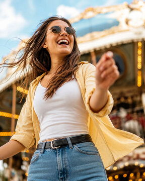 Joyful young woman laughing beside a brightly lit amusement ride, photographed from a low angle with energetic movement and soft carnival bokeh behind her.