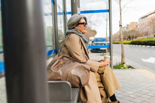 Woman with coat and scarf sitting at urban bus stop holding coffee